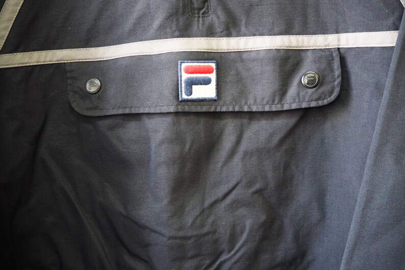 Op de afbeelding: Close-up van een donker marineblauwe jas met een zak en twee zilveren knopen. De zak heeft een rechthoekige patch met het Fila-logo in rood, wit en blauw. Een lichtbeige streep loopt over de borst.