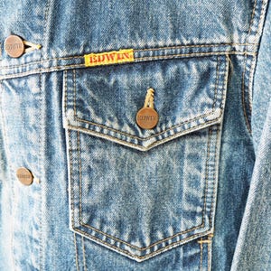 Puede incluir: Primer plano de una chaqueta vaquera con un lavado azul claro. La chaqueta tiene dos bolsillos en el pecho con cierre de bot&oacute;n y una peque&ntilde;a etiqueta amarilla con la marca "EDWIN". La chaqueta tiene botones color cobre.