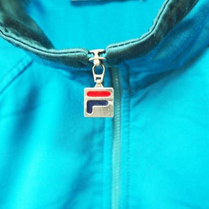 May include: Close-up of a turquoise athletic jacket with a black and teal collar and zipper. The zipper pull features a silver square with a red and blue logo. The jacket is made of a smooth, shiny material.