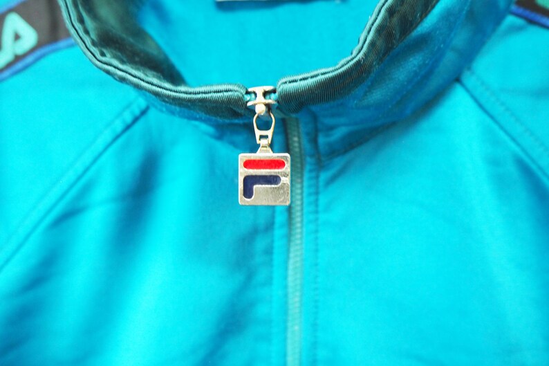 May include: Close-up of a turquoise athletic jacket with a black and teal collar and zipper. The zipper pull features a silver square with a red and blue logo. The jacket is made of a smooth, shiny material.