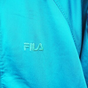 May include: A turquoise athletic jacket with a zipper closure and the embroidered brand name "FILA" in a matching turquoise thread. The sleeve features a black stripe with the brand name and logo in pink, blue, and green.