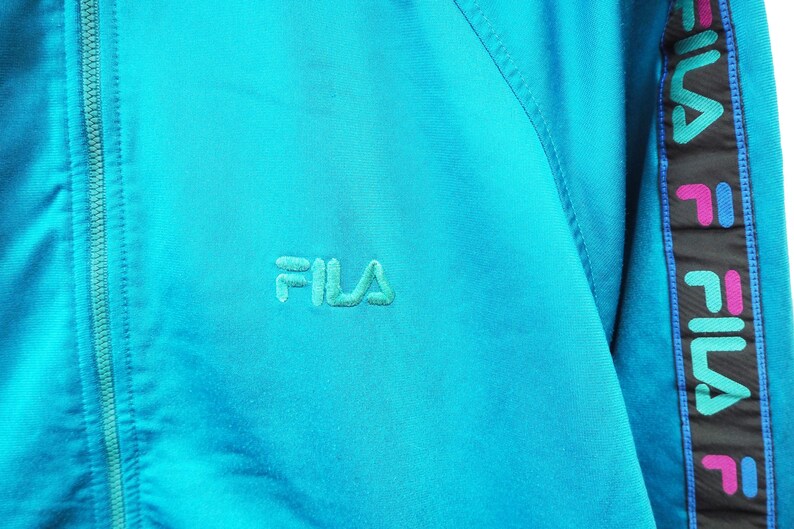 May include: A turquoise athletic jacket with a zipper closure and the embroidered brand name "FILA" in a matching turquoise thread. The sleeve features a black stripe with the brand name and logo in pink, blue, and green.