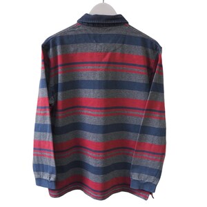 Puede incluir: Una camisa de manga larga con cuello y un estampado de rayas en rojo, gris y azul. La camisa est&aacute; colgada de una percha blanca sobre un fondo blanco.