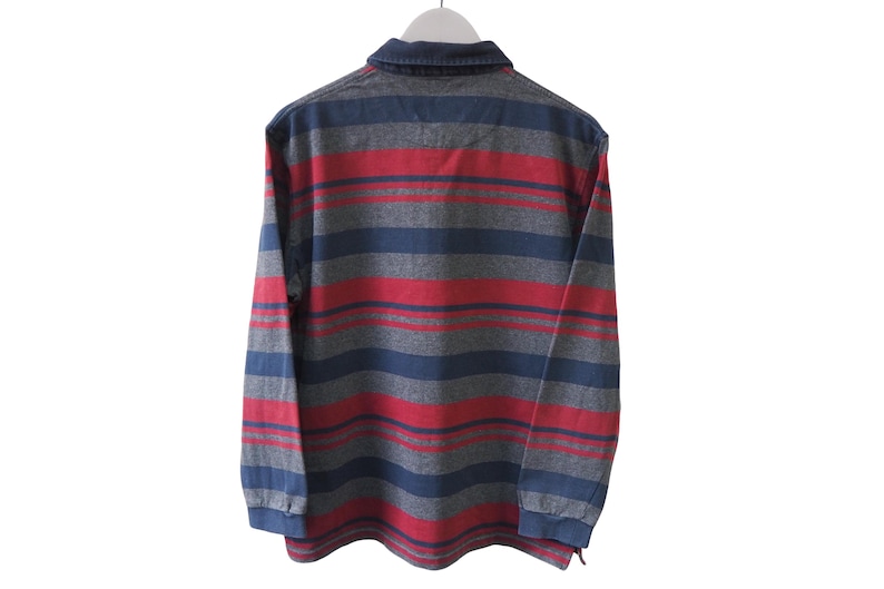 Puede incluir: Una camisa de manga larga con cuello y un estampado de rayas en rojo, gris y azul. La camisa est&aacute; colgada de una percha blanca sobre un fondo blanco.