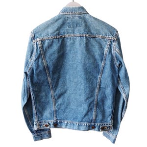 Puede incluir: Chaqueta vaquera azul claro, vista desde atr&aacute;s, colgada de una percha blanca. La chaqueta tiene un dise&ntilde;o cl&aacute;sico con cuello, botones y bolsillos. El tejido vaquero tiene un aspecto descolorido.