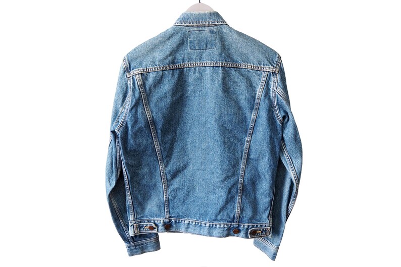 Puede incluir: Chaqueta vaquera azul claro, vista desde atr&aacute;s, colgada de una percha blanca. La chaqueta tiene un dise&ntilde;o cl&aacute;sico con cuello, botones y bolsillos. El tejido vaquero tiene un aspecto descolorido.