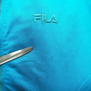 May include: Close-up of a turquoise blue athletic jacket with the embroidered brand name "FILA" in a matching color. A pair of scissors with pink handles is in the foreground, partially obscuring the jacket's fabric.