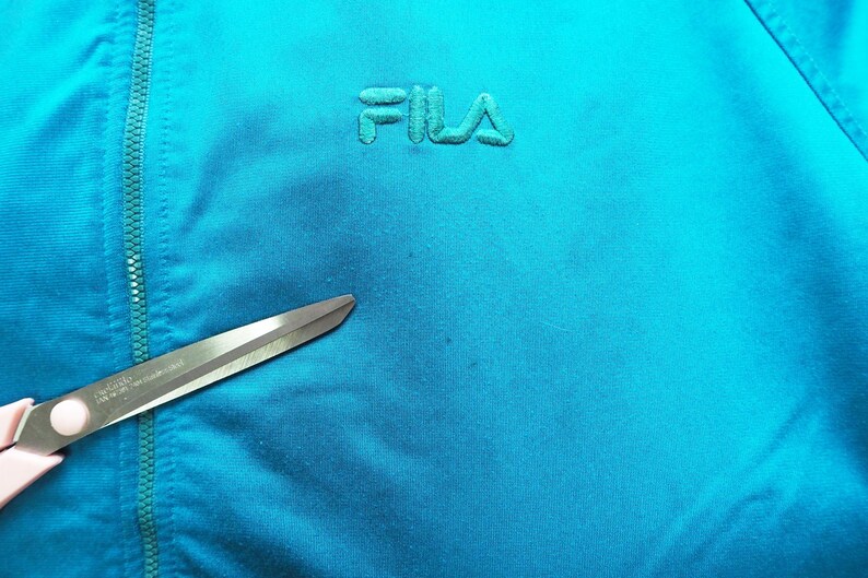 May include: Close-up of a turquoise blue athletic jacket with the embroidered brand name "FILA" in a matching color. A pair of scissors with pink handles is in the foreground, partially obscuring the jacket's fabric.