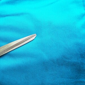 May include: Close-up of teal fabric with a zipper and a pair of scissors. The scissors are silver and green, and the fabric has a smooth texture. The zipper is teal and runs vertically.