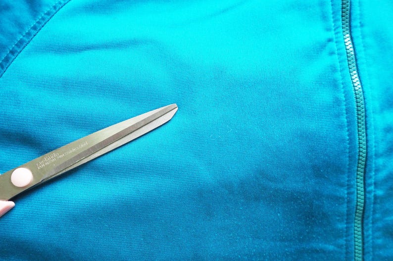 May include: Close-up of teal fabric with a zipper and a pair of scissors. The scissors are silver and green, and the fabric has a smooth texture. The zipper is teal and runs vertically.