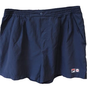 May include: Navy blue athletic shorts with an elastic waistband and a button closure. The shorts have two side pockets and a small logo patch on the lower right leg. These shorts are suitable for sports or casual wear.