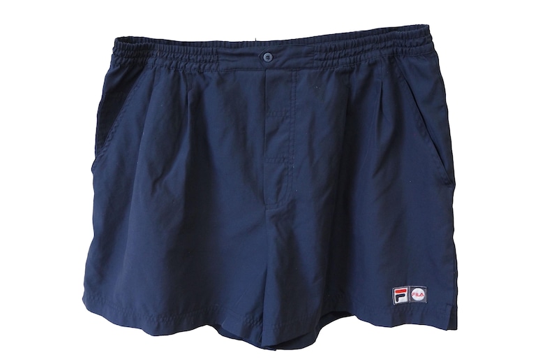 May include: Navy blue athletic shorts with an elastic waistband and a button closure. The shorts have two side pockets and a small logo patch on the lower right leg. These shorts are suitable for sports or casual wear.