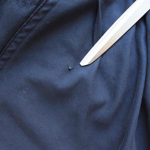May include: Close-up of navy blue fabric with a small imperfection. A white object, possibly a ruler or a straight edge, is placed diagonally across the fabric, pointing towards the imperfection. The fabric has visible stitching.