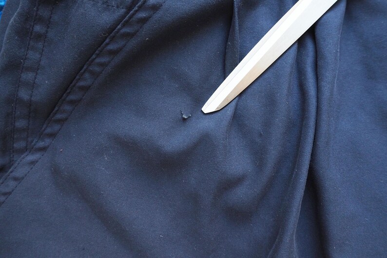 May include: Close-up of navy blue fabric with a small imperfection. A white object, possibly a ruler or a straight edge, is placed diagonally across the fabric, pointing towards the imperfection. The fabric has visible stitching.