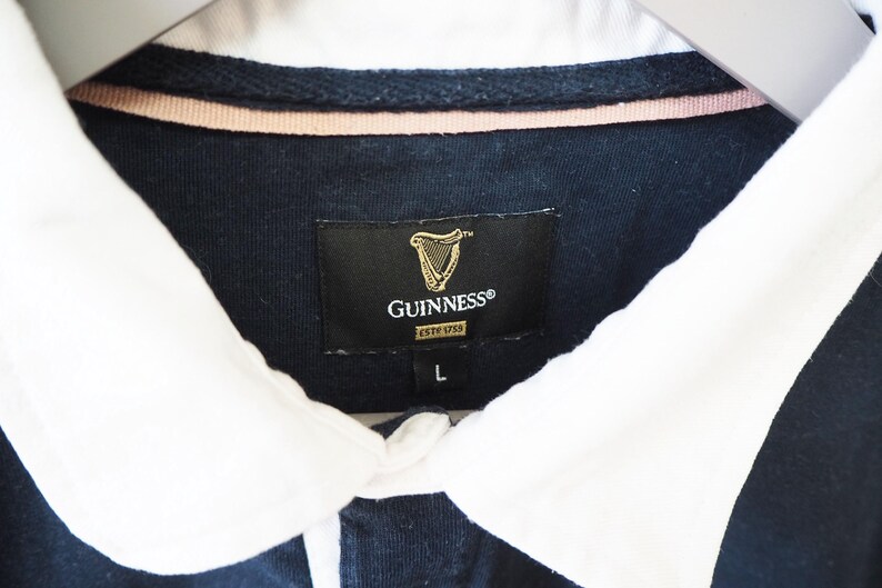 May include: Close-up of a navy blue rugby shirt with a white collar. The shirt features a Guinness label with a gold harp logo and the text "GUINNESS ESTD 1759". The size "L" is visible.