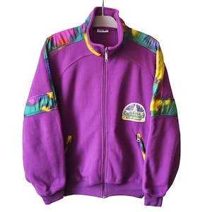 Peut inclure: Veste polaire violette avec fermeture éclair intégrale et col montant. La veste présente des accents colorés sur les épaules et un écusson sur la poitrine avec du texte. Deux poches zippées sont visibles.