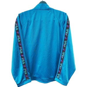 May include: A turquoise Fila track jacket with a high collar. The sleeves feature a black band with the Fila logo in a repeating pattern of pink, purple, and green. The jacket is hanging on a black hanger.