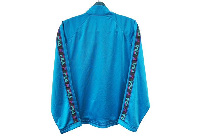 May include: A turquoise Fila track jacket with a high collar. The sleeves feature a black band with the Fila logo in a repeating pattern of pink, purple, and green. The jacket is hanging on a black hanger.