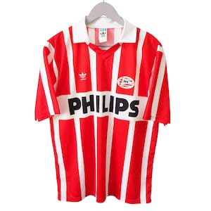 Op de afbeelding: Rood en wit gestreept voetbalshirt met het woord "PHILIPS" in zwart over de voorkant. Het shirt heeft een witte kraag en het Adidas-logo. Het shirt hangt aan een grijze hanger.