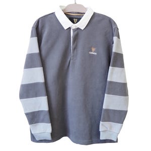 May include: A gray long-sleeve rugby shirt with white and gray striped sleeves. The shirt has a white collar and a small Guinness logo on the chest. The shirt is hanging on a hanger.