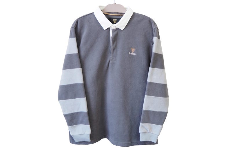 May include: A gray long-sleeve rugby shirt with white and gray striped sleeves. The shirt has a white collar and a small Guinness logo on the chest. The shirt is hanging on a hanger.