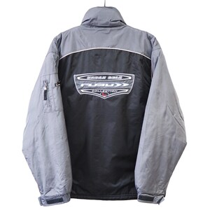 May include: A grey and black Fubu Collection jacket. The jacket has a high collar, a zip closure, and a logo on the back. The sleeves are grey, and the body is black with a grey panel across the shoulders.