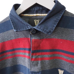 Puede incluir: Una camiseta de rugby de manga larga con rayas horizontales en azul marino, gris y rojo. La camiseta tiene un cuello abotonado y un peque&ntilde;o logotipo de Guinness cerca del dobladillo. El cuello es de un tono azul m&aacute;s oscuro.