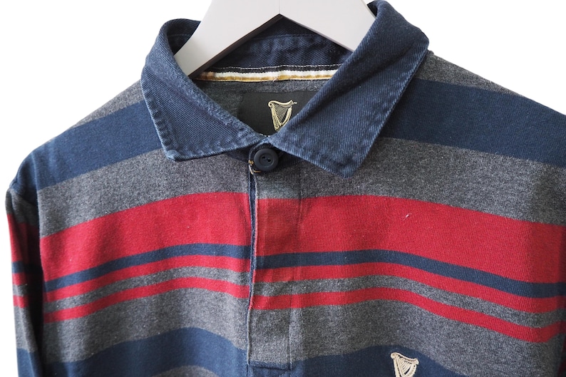 Puede incluir: Una camiseta de rugby de manga larga con rayas horizontales en azul marino, gris y rojo. La camiseta tiene un cuello abotonado y un peque&ntilde;o logotipo de Guinness cerca del dobladillo. El cuello es de un tono azul m&aacute;s oscuro.