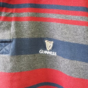 Puede incluir: Primer plano de una camiseta de rugby a rayas. La camiseta presenta rayas horizontales alternas rojas, grises y azul marino. Una banda azul marino en el pecho muestra el logotipo de Guinness en blanco.