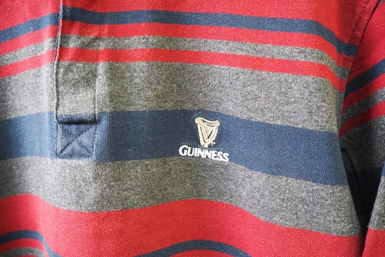 Puede incluir: Primer plano de una camiseta de rugby a rayas. La camiseta presenta rayas horizontales alternas rojas, grises y azul marino. Una banda azul marino en el pecho muestra el logotipo de Guinness en blanco.