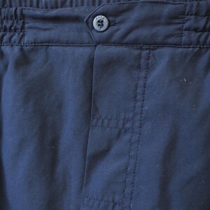 May include: Close-up of navy blue fabric, likely part of a garment. The image highlights the waistband with elastic gathering, a button, and vertical stitching details. The button has a small, circular design.