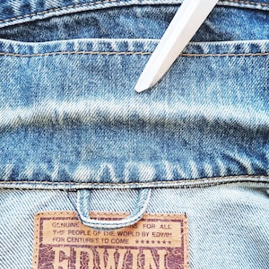 Puede incluir: Primer plano del cuello de una chaqueta vaquera azul descolorida. La chaqueta tiene costuras visibles y un parche de cuero marr&oacute;n con el texto "GENUINE THE PEOPLE OF THE WORLD BY EDWIN FOR CENTURIES TO COME". Un objeto blanco est&aacute; colocado sobre la chaqueta.
