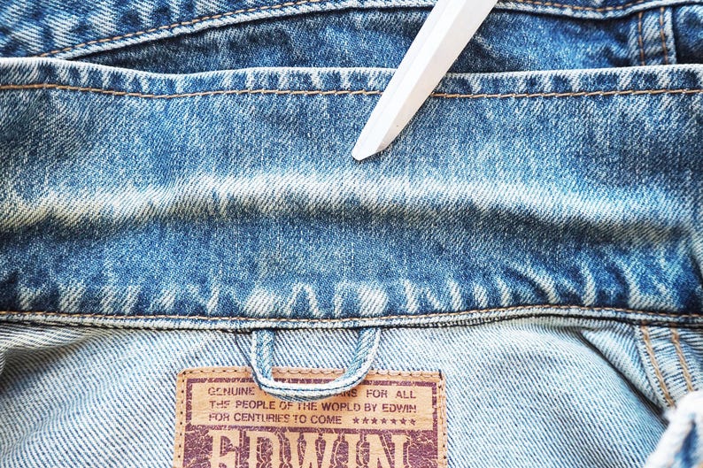 Puede incluir: Primer plano del cuello de una chaqueta vaquera azul descolorida. La chaqueta tiene costuras visibles y un parche de cuero marr&oacute;n con el texto "GENUINE THE PEOPLE OF THE WORLD BY EDWIN FOR CENTURIES TO COME". Un objeto blanco est&aacute; colocado sobre la chaqueta.