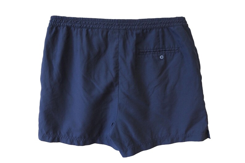 May include: Navy blue shorts with an elastic waistband and a single back pocket. The shorts are made of a lightweight fabric, ideal for warm weather. The back pocket has a button closure. The shorts are shown against a white background.