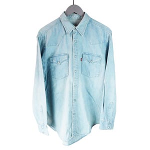May include: A light blue denim long-sleeve button-down shirt featuring a pointed collar, two chest pockets with snap closures, and a small red brand tag on the left pocket. The shirt has a faded wash and metal snap buttons down the front placket.
