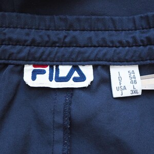 May include: Close-up of navy blue athletic pants with a stitched waistband. The Fila logo is visible on a white tag, along with size information. The pants are made of a smooth, possibly synthetic fabric.