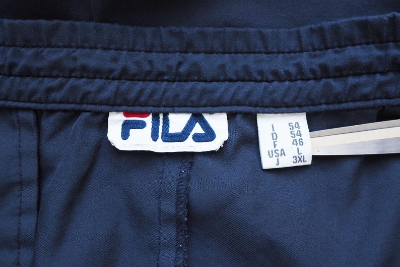 May include: Close-up of navy blue athletic pants with a stitched waistband. The Fila logo is visible on a white tag, along with size information. The pants are made of a smooth, possibly synthetic fabric.