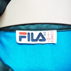 May include: Close-up of a teal and turquoise athletic jacket. The Fila logo is visible on a white tag, with red and blue accents. The tag also includes size information. The jacket's interior is turquoise, and the exterior is teal.