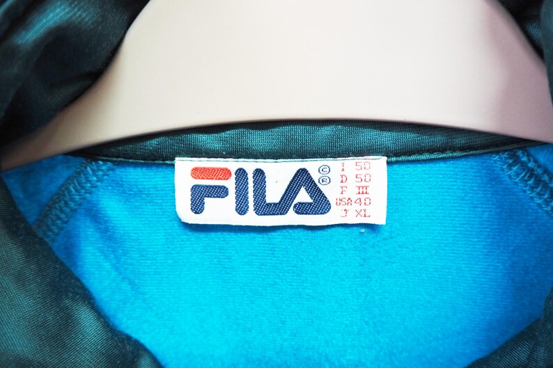 May include: Close-up of a teal and turquoise athletic jacket. The Fila logo is visible on a white tag, with red and blue accents. The tag also includes size information. The jacket's interior is turquoise, and the exterior is teal.