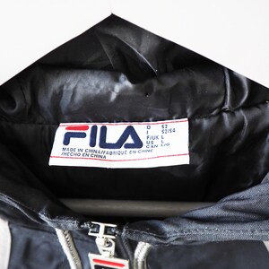 Op de afbeelding: Close-up van een zwarte FILA-jas met een wit en rood logo. De jas heeft een rits en witte strepen. Het label luidt "MADE IN CHINA". De jas hangt aan een witte hanger.