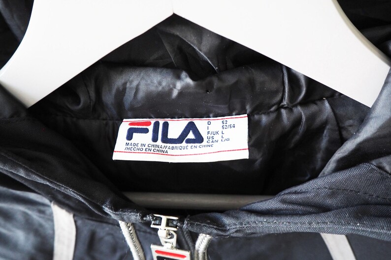 Op de afbeelding: Close-up van een zwarte FILA-jas met een wit en rood logo. De jas heeft een rits en witte strepen. Het label luidt "MADE IN CHINA". De jas hangt aan een witte hanger.