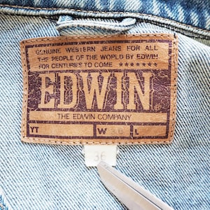 Puede incluir: Primer plano de una chaqueta vaquera con una etiqueta de cuero marr&oacute;n. La etiqueta dice "GENUINE WESTERN JEANS FOR ALL THE PEOPLE OF THE WORLD BY EDWIN" y presenta la marca "EDWIN". La etiqueta de talla muestra la talla 36.
