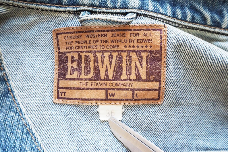 Puede incluir: Primer plano de una chaqueta vaquera con una etiqueta de cuero marr&oacute;n. La etiqueta dice "GENUINE WESTERN JEANS FOR ALL THE PEOPLE OF THE WORLD BY EDWIN" y presenta la marca "EDWIN". La etiqueta de talla muestra la talla 36.