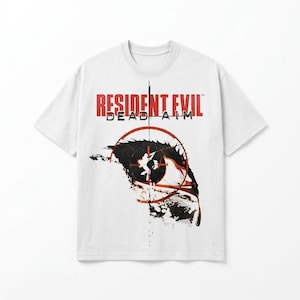 Puede incluir: Camiseta blanca con el texto "RESIDENT EVIL DEAD AIM" en rojo. El diseño incluye una diana sobre un ojo estilizado, con detalles en blanco y negro. Una camiseta clásica para los fans del videojuego.