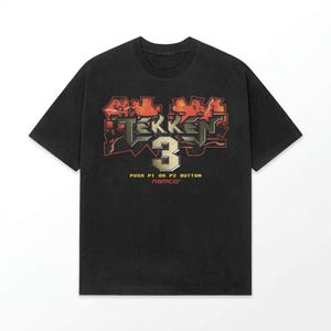 Può includere: T-shirt nera con il logo Tekken 3 in rosso, arancione e argento. La grafica include il numero 3 e il testo "PUSH P1 OR P2 BUTTON" e "namco". La maglietta ha maniche corte e girocollo.