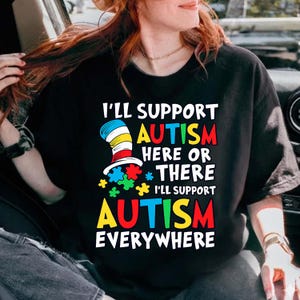 Op de afbeelding: Zwart t-shirt met de tekst "I'll Support Autism Here or There, I'll Support Autism Everywhere." Het ontwerp bevat een kleurrijke hoed en puzzelstukjes, met het woord "Autism" in rood, geel en blauw.