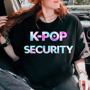 Peut inclure: T-shirt noir à col rond avec l'inscription "K-POP SECURITY" dans une police colorée sur le thème de la galaxie. Le motif présente des teintes bleues, roses et violettes, avec une étoile au centre du "P" de "K-POP".