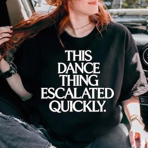 Puede incluir: Camiseta negra de cuello redondo con el texto blanco "THIS DANCE THING ESCALATED QUICKLY." La camiseta es de color negro liso y parece estar hecha de un material suave. El texto está en negrita, sin serifa.