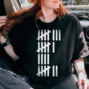 Peut inclure: T-shirt noir avec un motif de marques de pointage blanches. Le motif est composé de lignes verticales avec des barres obliques, représentant un compte. C'est un vêtement décontracté pour tous les jours.