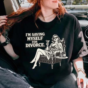 Op de afbeelding: Zwart t-shirt met de tekst "I'M SAVING MYSELF FOR DIVORCE." De afbeelding toont een vrouw in een badjas, zittend in een stoel, met een cocktail in haar hand. Op een tafeltje staat een fles en een glas.